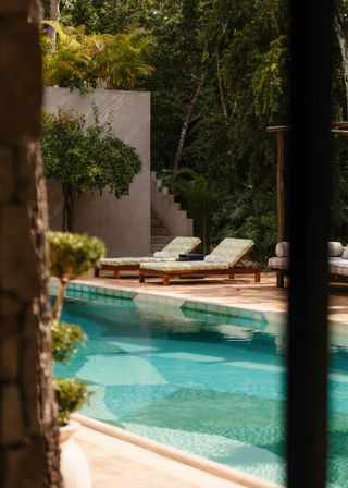 Turquoise pool at a tropical villa oasis with a wooden deck, two patterned sun loungers with rolled towels by concrete steps, surrounded by lush jungle foliage.