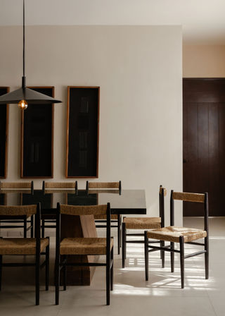 Sunlit minimalist dining room with dark wood table, woven-seat chairs, black pendant light and framed black wall art on neutral plaster walls