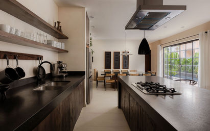 Open-plan modern kitchen with a large dark stone island and gas cooktop, floating wooden shelves with glassware, stainless refrigerator, black pendant lights, and a sunlit dining area by sliding glass doors with tropical plants visible.