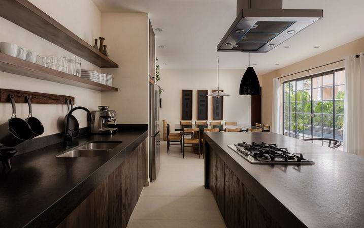 Open-plan modern kitchen with a large dark stone island and gas cooktop, floating wooden shelves with glassware, stainless refrigerator, black pendant lights, and a sunlit dining area by sliding glass doors with tropical plants visible.
