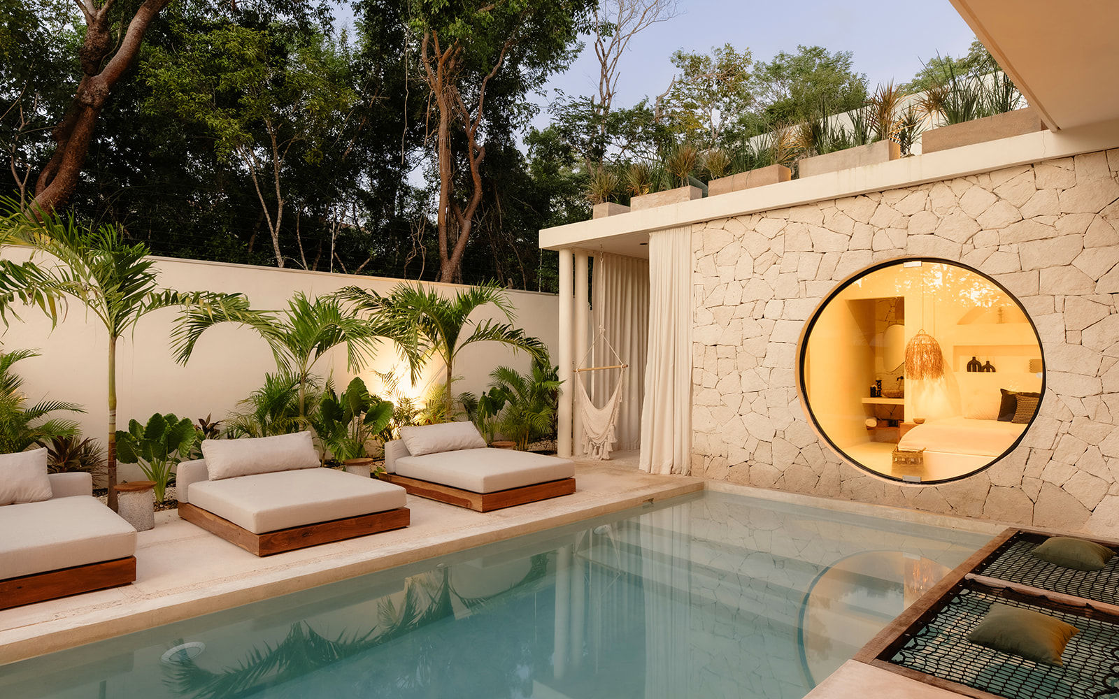 Modern tropical poolside patio with cushioned wooden daybeds, hammock and palms beside a calm rectangular pool; round stone window reveals a warm-lit bedroom.