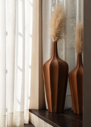 Sunlit boho window scene with two tall terracotta vases and fluffy pampas grass on a dark windowsill beside sheer white curtains.
