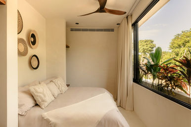 Sunlit modern minimalist bedroom with cream bedding and textured pillows, woven wall baskets, wooden ceiling fan, and a large window opening to lush tropical plants