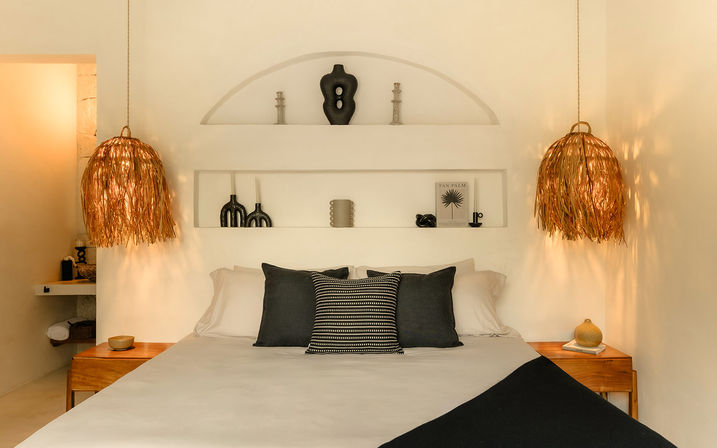 Cozy Mediterranean-inspired bedroom with white linens, black and striped accent pillows, straw-fringe pendant lamps over wooden nightstands, recessed wall niche with sculptural vases and candles, and warm ambient lighting.