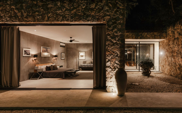 Warm-lit modern bedroom in a stone-clad villa with an open sliding wall onto a gravel courtyard at night