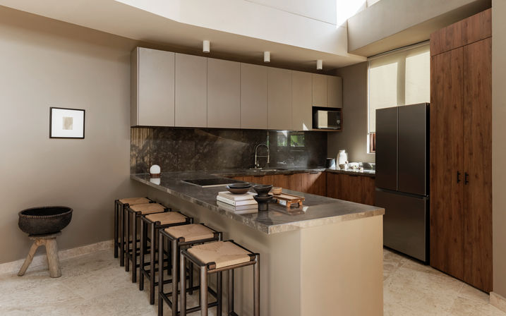 Sunlit modern kitchen interior with marble-topped island, row of woven-seat bar stools, matte wood cabinets, dark stone backsplash, black refrigerator and skylight