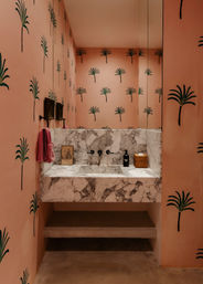 Stylish pink powder room with green palm-print walls, marble floating vanity and integrated sink, black wall-mounted faucets, mirror, pink towel and wicker soap basket.