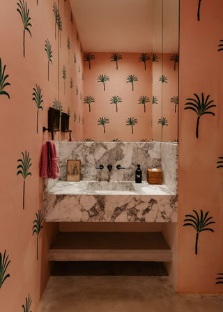 Stylish pink powder room with green palm-print walls, marble floating vanity and integrated sink, black wall-mounted faucets, mirror, pink towel and wicker soap basket.