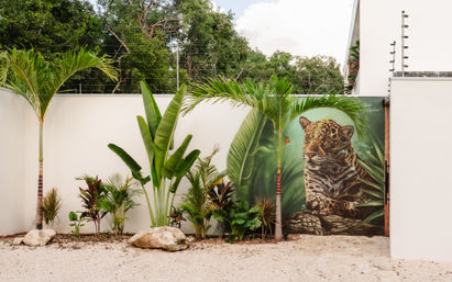Outdoor tropical courtyard with palm trees and lush plants along a white wall showcasing a vibrant jaguar street-art mural framed by oversized green leaves.