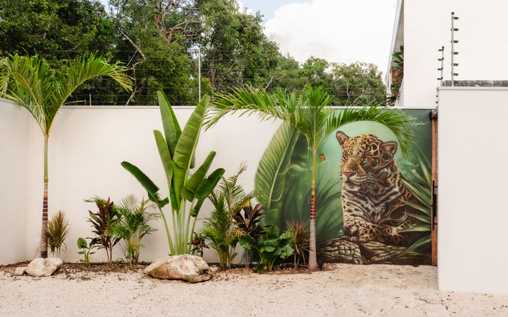 Outdoor tropical courtyard with palm trees and lush plants along a white wall showcasing a vibrant jaguar street-art mural framed by oversized green leaves.