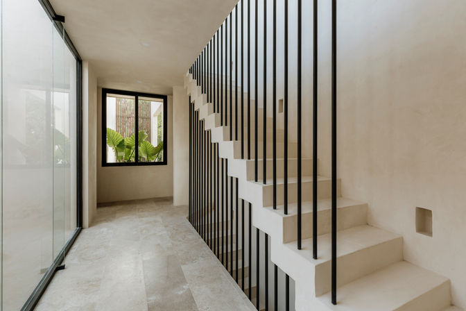 Minimalist modern hallway with white concrete staircase and black vertical balusters, stone tile flooring and a large window revealing lush tropical plants.