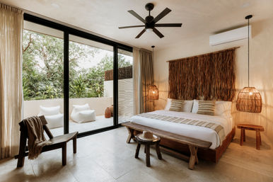 Sunlit tropical boho bedroom with king bed, woven rattan pendant lights, ceiling fan and textured wall hanging; floor-to-ceiling sliding glass doors open to a private patio with greenery and lounge poufs, neutral tones and wooden accents.