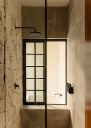 Sunlit modern shower with exposed stone wall, black rain showerhead, industrial grid window and neutral stone tiles, spa-like minimalist bathroom