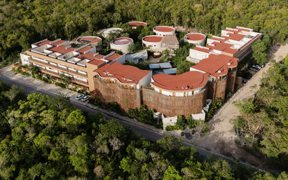 Aerial view of a modern eco-hotel complex with terracotta red roofs and circular rooftop villas nestled in dense tropical forest, curved wooden facades surrounding inner courtyards and pools, with a dirt road and parked cars visible.