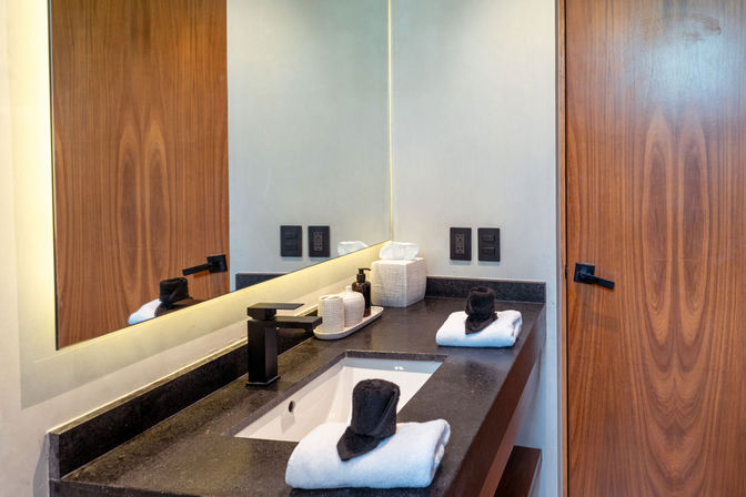 Sleek modern hotel bathroom vanity with black stone countertop, undermount sink and matte-black faucet, folded white towels topped with rolled black hand towels, woven tissue box and soap dispenser, large mirror and wooden door.