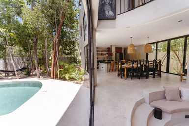 Open-plan tropical villa showing poolside patio, sliding glass walls to dining area with wooden table, black chairs and rattan pendant lights, adjacent kitchen island with bar stools, curved built-in sofa and lush trees with a hammock outside.