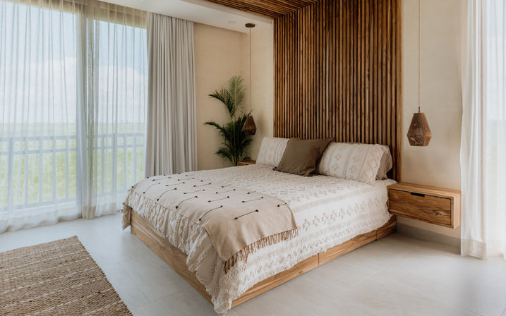 Sunlit modern boho bedroom interior with wooden slatted accent wall, king bed with textured cream bedding, hanging pendant lights, potted plant and sheer curtains opening to a balcony view.