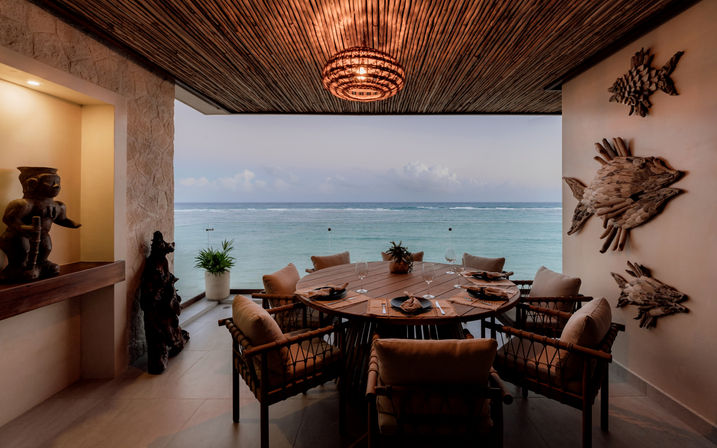 Covered coastal dining terrace with a round wooden table set for six, cushioned wicker chairs, carved fish wall art and sculptures, a woven pendant light, potted plants and a sweeping ocean view.