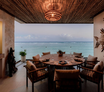 Covered coastal dining terrace with a round wooden table set for six, cushioned wicker chairs, carved fish wall art and sculptures, a woven pendant light, potted plants and a sweeping ocean view.