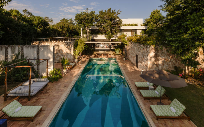 Long rectangular turquoise lap pool at a modern tropical villa, flanked by stone terraces, striped daybeds and green-patterned sun loungers with umbrella, surrounded by lush trees and a white contemporary house in golden-hour light.