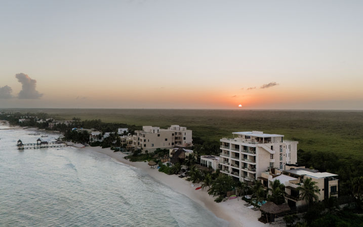 Aerial view of a tropical beachfront at sunset with white-sand shore, palm trees, low-rise beachfront condos, a wooden pier stretching into turquoise water, and the sun dipping over a green coastal plain.