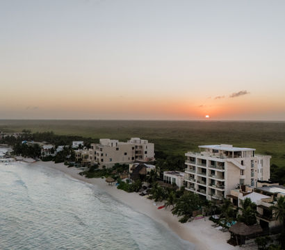 Aerial view of a tropical beachfront at sunset with white-sand shore, palm trees, low-rise beachfront condos, a wooden pier stretching into turquoise water, and the sun dipping over a green coastal plain.