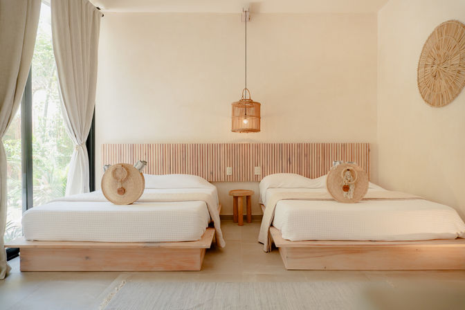 Bright boho resort bedroom with two low wooden beds, white bedding, round straw accent pillows, rattan pendant light and garden-view floor-to-ceiling window