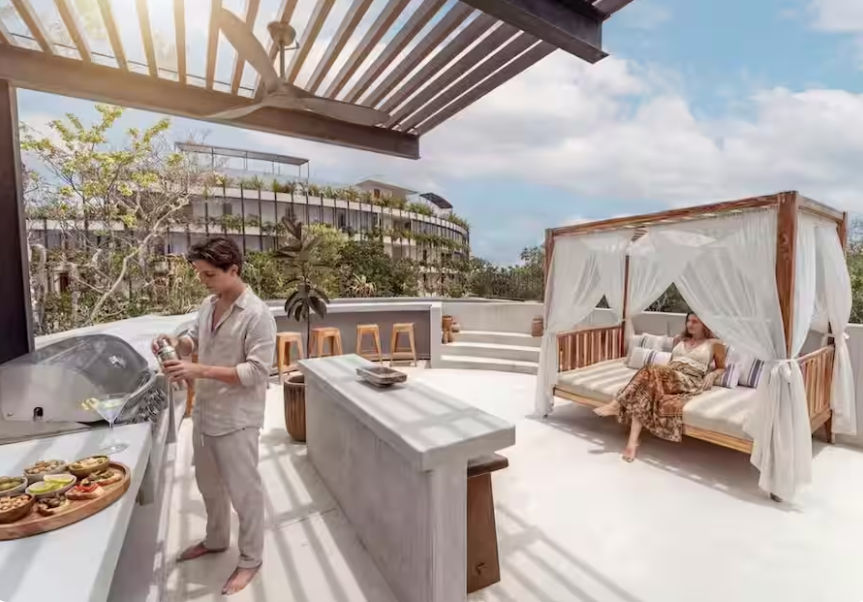 Sunny tropical rooftop terrace at a resort with a pergola‑shaded outdoor kitchen and bar, a guest mixing a cocktail at the counter and another relaxing on a draped daybed cabana.