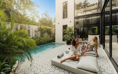 Two friends lounging by a turquoise plunge pool at a modern glass‑walled villa patio — patterned geometric tile, lush tropical plants, sunlit relaxing vibe with a pink cocktail.