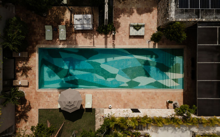 Aerial bird’s-eye view of a long rectangular pool with blue-green geometric mosaic tiles, terracotta patio, lounge chairs, umbrella, and tropical landscaping.