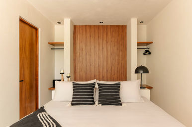 Cozy modern minimalist bedroom with white bedding, two black-and-white striped pillows, wood slatted accent wall, floating shelves and a black bedside lamp.