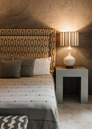 Cozy modern bedroom interior with geometric patterned headboard, neutral bedding and pillows, and a striped lampshade on a concrete bedside table.
