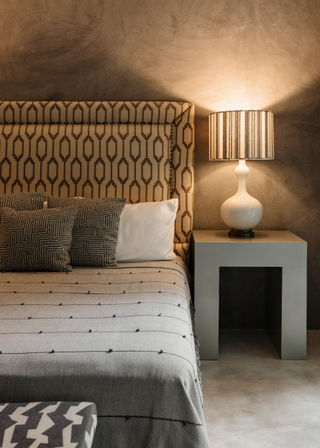 Cozy modern bedroom interior with geometric patterned headboard, neutral bedding and pillows, and a striped lampshade on a concrete bedside table.