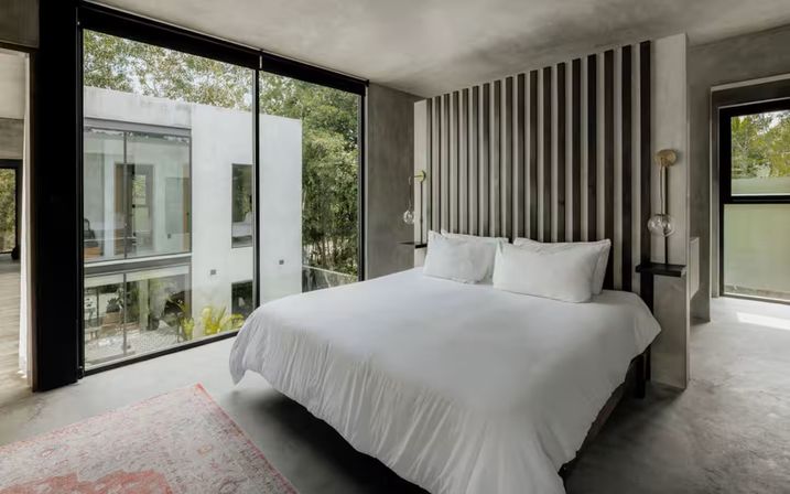 Minimalist modern bedroom with white bedding, vertical slatted headboard, glass pendant lights and floor-to-ceiling window overlooking trees and a courtyard.