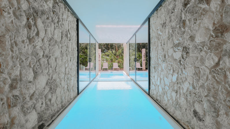 Turquoise indoor-outdoor pool corridor framed by rough stone walls and sliding glass doors, opening to a pool deck with lounge chairs and lush tropical greenery.