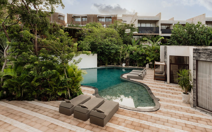 Curved turquoise pool at a tropical luxury villa resort, tiled terrace with cushioned sun loungers, lush palms and modern guest rooms in the background.