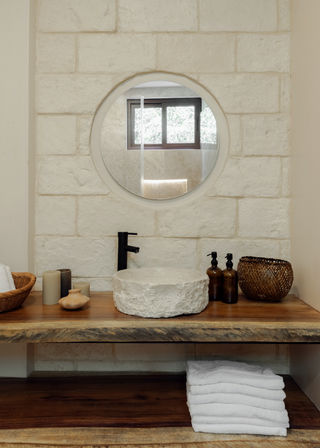 Spa-style rustic bathroom vanity with round mirror, rough stone vessel sink and matte black faucet on a natural wood countertop, amber soap dispensers, woven basket, candles and folded white towels against a pale stone wall