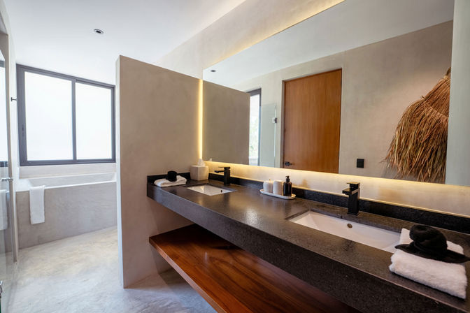Modern minimalist bathroom with a long black stone double vanity, matte black faucets, backlit mirror, wooden shelf, folded towels and a frosted window providing natural light.