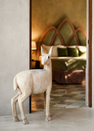 Playful carved wooden deer sculpture standing in a doorway, leading to a cozy bedroom with an arched headboard, green velvet pillows and pastel patterned tile floor.