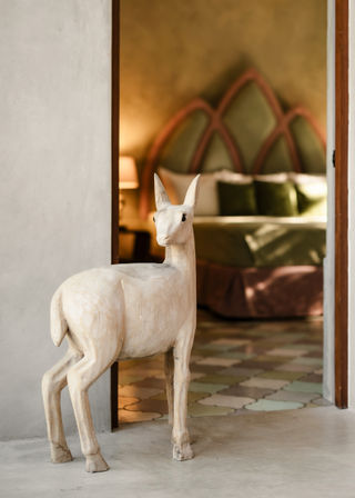 Playful carved wooden deer sculpture standing in a doorway, leading to a cozy bedroom with an arched headboard, green velvet pillows and pastel patterned tile floor.