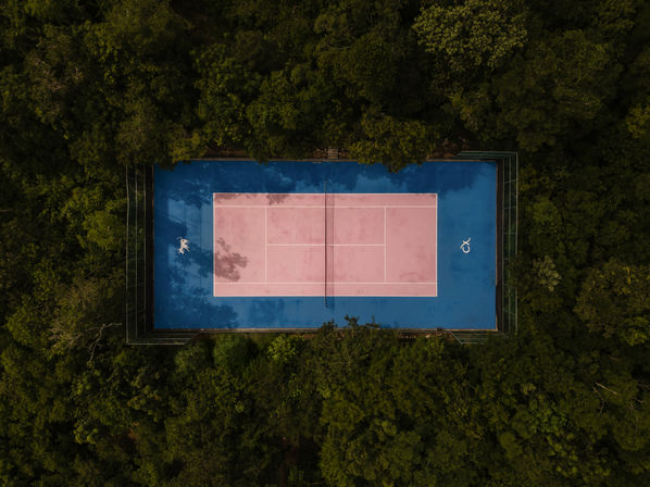 Aerial drone view of a pastel pink tennis court with blue outer area nestled inside dense green forest, overhead composition with a lone person on the court.