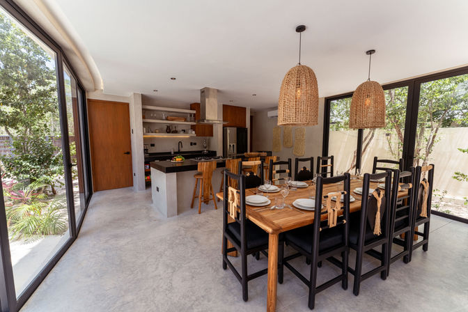 Open-plan modern dining room and kitchen with a long wooden table set for dinner, black chairs with rope accents, woven rattan pendant lights, concrete floor and floor-to-ceiling windows overlooking a lush garden.