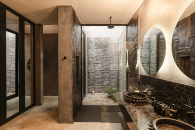 Modern spa-style bathroom with an indoor courtyard shower and pebble floor, textured gray tile walls, black stone double sinks with round backlit mirrors, and concrete-industrial finishes.
