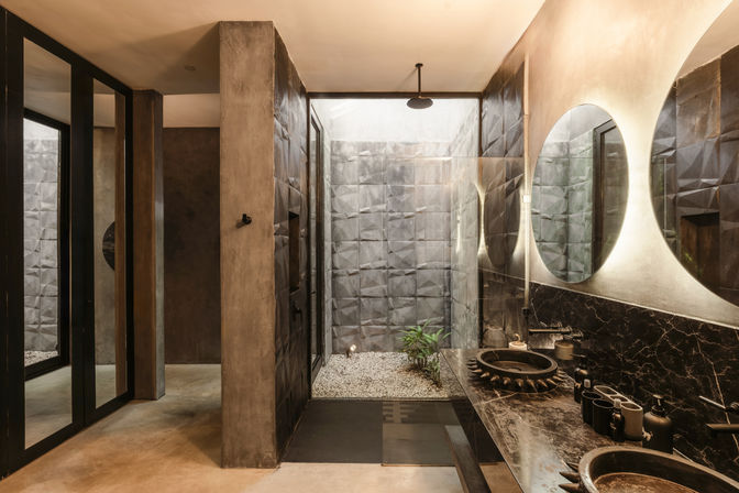 Modern spa-style bathroom with an indoor courtyard shower and pebble floor, textured gray tile walls, black stone double sinks with round backlit mirrors, and concrete-industrial finishes.