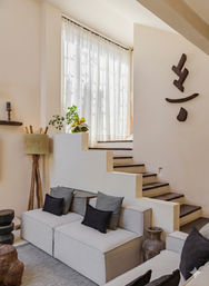 Sunlit modern neutral living room with beige modular sofa and black cushions, stepped plaster partition leading to dark wooden stairs, sheer floor-to-ceiling curtains, potted plants, rustic wood wall art and textured accessories.