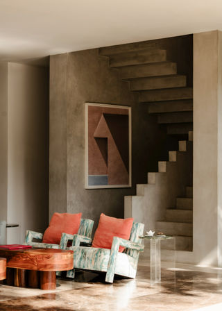 Sunlit modern living room interior with sculptural concrete staircase, polished floor, two turquoise patterned armchairs with coral cushions, round wooden coffee table, clear acrylic side table and geometric wall art.
