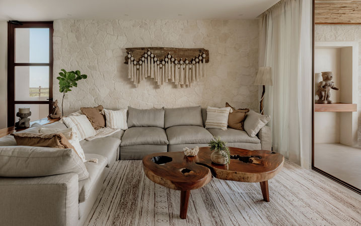 Sunlit neutral boho living room with a large linen sectional, textured stone accent wall and macramé wall hanging, live-edge wooden coffee table on a patterned rug, sheer curtains and potted fiddle-leaf fig for a natural, coastal-inspired look.