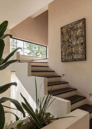 Sunlit modern interior staircase with dark wood treads, large multi-pane window, potted green plants, and abstract wall art in warm neutral tones