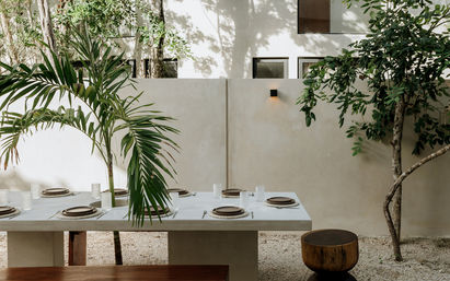 Minimalist outdoor dining patio in a tropical courtyard: modern concrete table set for six with woven chargers and glassware, palm fronds and leafy trees against a neutral stucco wall and gravel floor.