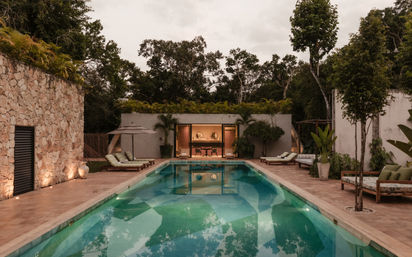 Tropical resort-style villa with a long illuminated outdoor swimming pool, cushioned lounge chairs, umbrellas and a warm-lit open pavilion set among lush trees at dusk.